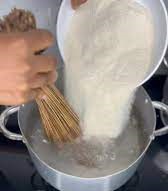 turning semo from ceramic bowl, into a boiling pot of water on a gas stove with a hand holding a broom as spatula, turning the semo in turned into the pot